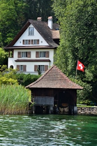 A large, traditional Swiss-style house with a steep roof and multiple windows is surrounded by lush greenery. In the foreground, there is a wooden boathouse next to calm water. A Swiss flag is visible near the boathouse, indicating the location.