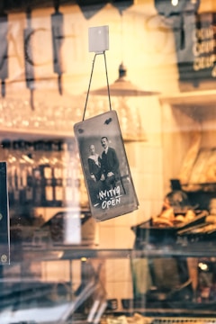 A vintage-style photograph resembling an old portrait hangs in a shop window. The sign on the photograph reads 'Always Open' in an artistic font. The background shows a warm-toned interior of a café or bakery with shelves likely filled with bread or pastries. Some reflections on the glass can be seen, creating a nostalgic ambiance.