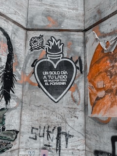 A piece of street art featuring a stylized illustration of a heart with flames on top and a flower to the upper left. The heart contains the text 'UN SOLO DÍA A TU LADO ME VALE POR TODO EL PORVENIR.' The wall is covered with faded graffiti and layers of orange, black, and gray paint, with some peeling sections.