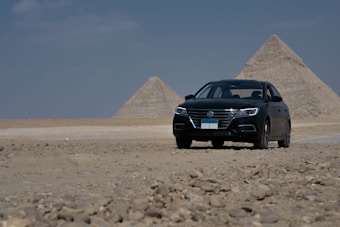 A black car is parked on a desert landscape, with two pyramids in the background. The scene suggests a mix of modern and ancient elements, set against a clear blue sky.
