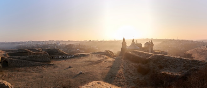 A panoramic view of a landscape featuring a historic fortress with prominent towers and walls, set against a sunrise or sunset sky. The scene is infused with soft, warm light creating long shadows on the ground. The surrounding area appears natural and rugged, with distant buildings in the background forming a cityscape.