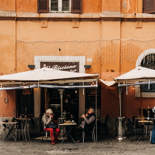 An outdoor cafe with a vibrant orange facade features two patrons seated under large white umbrellas. One person is wearing a gray coat and red pants, while the other is in dark attire. The cafe sign reads 'Al Biscione Cafe,' and decorative lights hang above the entrance. Two tables have small potted plants, and a waiter can be seen inside the cafe wearing a mask.