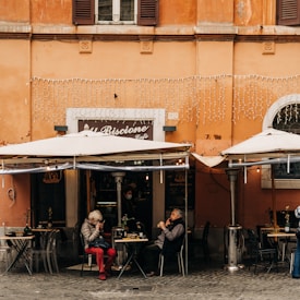 An outdoor cafe with a vibrant orange facade features two patrons seated under large white umbrellas. One person is wearing a gray coat and red pants, while the other is in dark attire. The cafe sign reads 'Al Biscione Cafe,' and decorative lights hang above the entrance. Two tables have small potted plants, and a waiter can be seen inside the cafe wearing a mask.