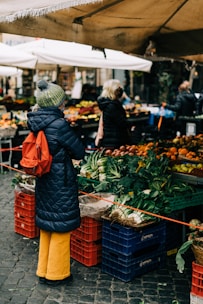 woman in black knit cap and orange coat standing in front of green vegetable display