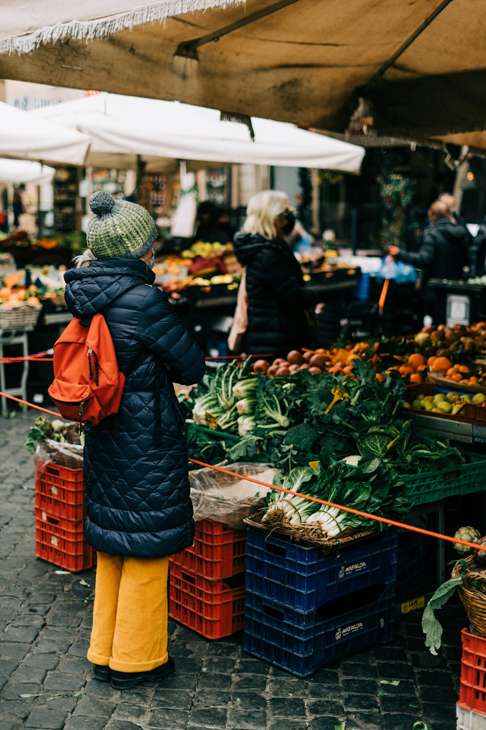 Femme en bonnet en tricot noir et manteau orange debout devant l’étalage de légumes verts