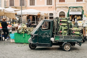 An outdoor market scene with a small green delivery vehicle parked beside stalls with fresh produce. The vehicle is loaded with crates of vegetables. Two people are engaged in market activities, one standing next to a stall filled with artichokes, while another person is in the background near a deli. The market is set against old European-style buildings, and awnings cover some of the stalls.
