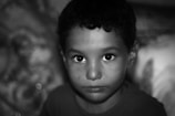 A close-up portrait of a young child with short curly hair, staring intently at the camera. The background is blurred, emphasizing the child's expression and wide eyes. The image is in black and white, adding a timeless and classic feel.