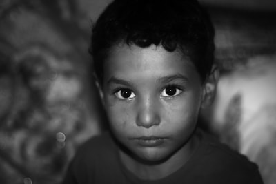A close-up portrait of a young child with short curly hair, staring intently at the camera. The background is blurred, emphasizing the child's expression and wide eyes. The image is in black and white, adding a timeless and classic feel.