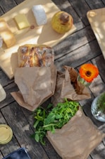 A rustic picnic setup on a wooden table featuring a variety of foods. Slices of cheese and a yellow pear rest on a wooden board. A loaf of bread is wrapped in brown paper, next to a bundle of fresh green leafy vegetables spilling out of another paper bag. An orange flower adds a pop of color to the scene.