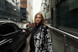 A young woman stands confidently on an urban street, framed by modern buildings. She wears a stylish black jacket adorned with white graphic text and symbols. The street is lined with parked cars, adding to the city vibe.