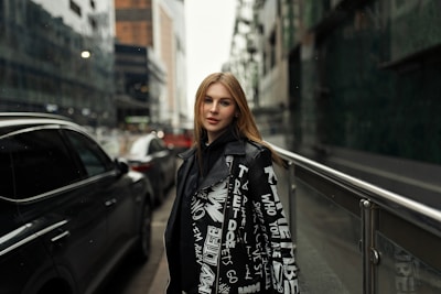 A young woman stands confidently on an urban street, framed by modern buildings. She wears a stylish black jacket adorned with white graphic text and symbols. The street is lined with parked cars, adding to the city vibe.
