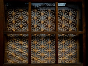 A wooden window with a geometric lattice design featuring intricate patterns of interlocking stars and triangles that form a decorative screen. The lattice is made of light-colored wood against a darker brown frame, and natural light creates subtle shadows within the grid.