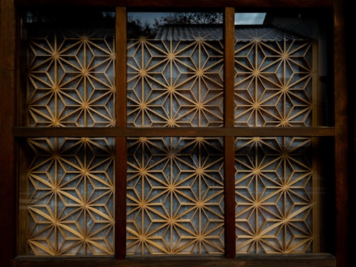 A wooden window with a geometric lattice design featuring intricate patterns of interlocking stars and triangles that form a decorative screen. The lattice is made of light-colored wood against a darker brown frame, and natural light creates subtle shadows within the grid.