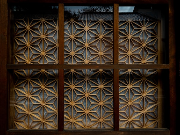 A wooden window with a geometric lattice design featuring intricate patterns of interlocking stars and triangles that form a decorative screen. The lattice is made of light-colored wood against a darker brown frame, and natural light creates subtle shadows within the grid.