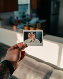 A hand with tattoos holds a Polaroid photo depicting a baby sitting in a high chair with food on the tray. The background shows a kitchen with blurred plants and a refrigerator.