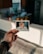 A hand with tattoos holds a Polaroid photo depicting a baby sitting in a high chair with food on the tray. The background shows a kitchen with blurred plants and a refrigerator.