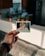 A hand with tattoos holds a Polaroid photo depicting a baby sitting in a high chair with food on the tray. The background shows a kitchen with blurred plants and a refrigerator.