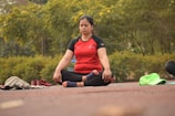 A person sits cross-legged on an outdoor mat with eyes closed, engaged in meditation. Surrounding them is a lush, green park with vibrant yellow flowers. Beside the individual are some personal items, including a pair of red shoes and some clothing.