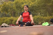 A person sits cross-legged on an outdoor mat with eyes closed, engaged in meditation. Surrounding them is a lush, green park with vibrant yellow flowers. Beside the individual are some personal items, including a pair of red shoes and some clothing.