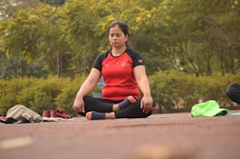 A person sits cross-legged on an outdoor mat with eyes closed, engaged in meditation. Surrounding them is a lush, green park with vibrant yellow flowers. Beside the individual are some personal items, including a pair of red shoes and some clothing.