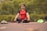 A person sits cross-legged on an outdoor mat with eyes closed, engaged in meditation. Surrounding them is a lush, green park with vibrant yellow flowers. Beside the individual are some personal items, including a pair of red shoes and some clothing.
