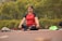A person sits cross-legged on an outdoor mat with eyes closed, engaged in meditation. Surrounding them is a lush, green park with vibrant yellow flowers. Beside the individual are some personal items, including a pair of red shoes and some clothing.