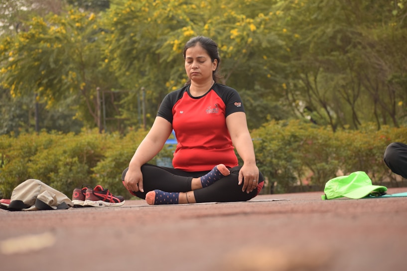 A person sits cross-legged on an outdoor mat with eyes closed, engaged in meditation. Surrounding them is a lush, green park with vibrant yellow flowers. Beside the individual are some personal items, including a pair of red shoes and some clothing.