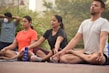 A group of people practicing yoga outdoors, sitting cross-legged with eyes closed. Each person appears to be focused and relaxed. There are trees in the background, indicating a peaceful and natural setting.