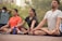 A group of people practicing yoga outdoors, sitting cross-legged with eyes closed. Each person appears to be focused and relaxed. There are trees in the background, indicating a peaceful and natural setting.
