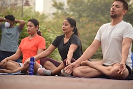 A group of people practicing yoga outdoors, sitting cross-legged with eyes closed. Each person appears to be focused and relaxed. There are trees in the background, indicating a peaceful and natural setting.