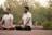 Two individuals are sitting cross-legged outdoors on a mat with their eyes closed, practicing meditation or yoga in a serene park setting. In the background, a bicycle is visible alongside lush greenery and trees, creating a calm atmosphere.