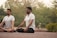 Two individuals are sitting cross-legged outdoors on a mat with their eyes closed, practicing meditation or yoga in a serene park setting. In the background, a bicycle is visible alongside lush greenery and trees, creating a calm atmosphere.