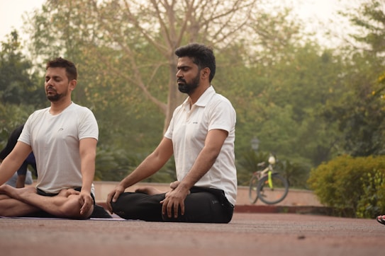Two individuals are sitting cross-legged outdoors on a mat with their eyes closed, practicing meditation or yoga in a serene park setting. In the background, a bicycle is visible alongside lush greenery and trees, creating a calm atmosphere.