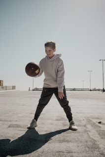 A young person is outdoors on a sunlit basketball court, engaged in dribbling a basketball. They are wearing a light hoodie and dark pants, and appear to be focused with a cheerful expression. The background reveals an empty, expansive space with clear blue sky and distant structures.