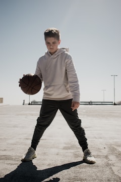 A young person stands in an open outdoor area holding a basketball, dressed in a light hoodie and dark sweatpants. The sky is clear, and the ground is a concrete surface, with the late afternoon sun casting a distinct shadow.