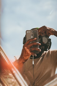 A person holding a smartphone is taking a mirror selfie. The person is wearing a beige shirt and has a large pair of headphones around their neck. The focus is on the hand and smartphone, with the face and background slightly out of focus. The scene has a warm, relaxed feel with natural lighting.