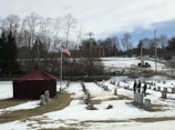 A snow-covered cemetery features rows of gravestones, a red tent, and two flagpoles with flags. Several people dressed in uniforms are standing near the gravestones. In the background, there are leafless trees, a brick building, and a cloudy sky.