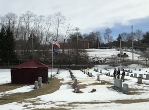 A snow-covered cemetery features rows of gravestones, a red tent, and two flagpoles with flags. Several people dressed in uniforms are standing near the gravestones. In the background, there are leafless trees, a brick building, and a cloudy sky.