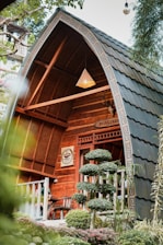 A quaint, wooden cabin with arched roof features intricate woodwork and a cozy seating area on the porch. Surrounding it are well-manicured shrubs, trees, and colorful plants, creating a serene, natural setting. A sign with a coffee cup symbol is mounted near the entrance, hinting at a cafe atmosphere. A hanging lamp adds warmth to the wooden structure.