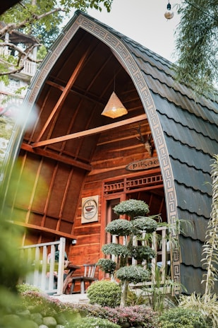 A quaint, wooden cabin with arched roof features intricate woodwork and a cozy seating area on the porch. Surrounding it are well-manicured shrubs, trees, and colorful plants, creating a serene, natural setting. A sign with a coffee cup symbol is mounted near the entrance, hinting at a cafe atmosphere. A hanging lamp adds warmth to the wooden structure.