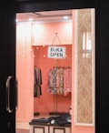 A view into a clothing store through a glass door with a sign that reads 'BUKA OPEN' hanging on it. Several stylish dresses and garments are visible on racks inside the store, against a soft pink interior with decorative patterns on the walls.