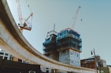 A construction site features prominently with two tall cranes working around partially constructed buildings clad in blue safety netting. An elevated roadway curves across the scene, while the sky is clear, and several brick commercial buildings are visible in the background.