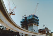 A construction site features prominently with two tall cranes working around partially constructed buildings clad in blue safety netting. An elevated roadway curves across the scene, while the sky is clear, and several brick commercial buildings are visible in the background.