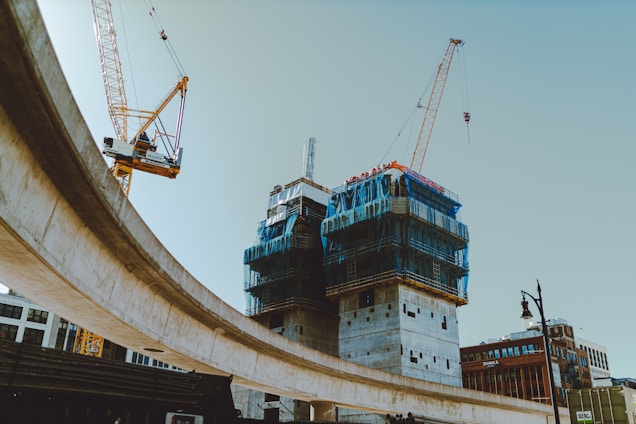 A construction site features prominently with two tall cranes working around partially constructed buildings clad in blue safety netting. An elevated roadway curves across the scene, while the sky is clear, and several brick commercial buildings are visible in the background.