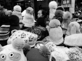 An assortment of knit hats with pom-poms displayed on a market stall. A person is partially visible among the hats, which are arranged in various styles and patterns.