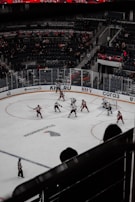 A hockey game in an indoor arena with players in red and white jerseys competing on the ice. The arena is partially filled with spectators, and banners are displayed in the stands. The scene captures the action mid-game with players and a referee on the ice.