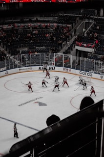 A hockey game in an indoor arena with players in red and white jerseys competing on the ice. The arena is partially filled with spectators, and banners are displayed in the stands. The scene captures the action mid-game with players and a referee on the ice.