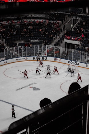 A hockey game in an indoor arena with players in red and white jerseys competing on the ice. The arena is partially filled with spectators, and banners are displayed in the stands. The scene captures the action mid-game with players and a referee on the ice.