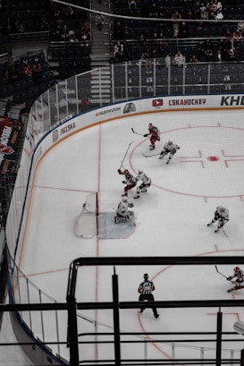 An ice hockey game is in progress, featuring players wearing red and white uniforms. The scene captures action around the goal area as players strive to control the puck. A referee is visible on the ice, observing the game closely. Spectators are seated in the background, watching the match.