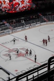 A hockey game is taking place on an indoor ice rink. Players, dressed in red and white uniforms, are gathered around the center ice area, marked with a large star logo. Several referees in black and white striped uniforms are also on the ice. Above the rink is a large digital scoreboard displaying red star graphics. The stands are partially filled with spectators.